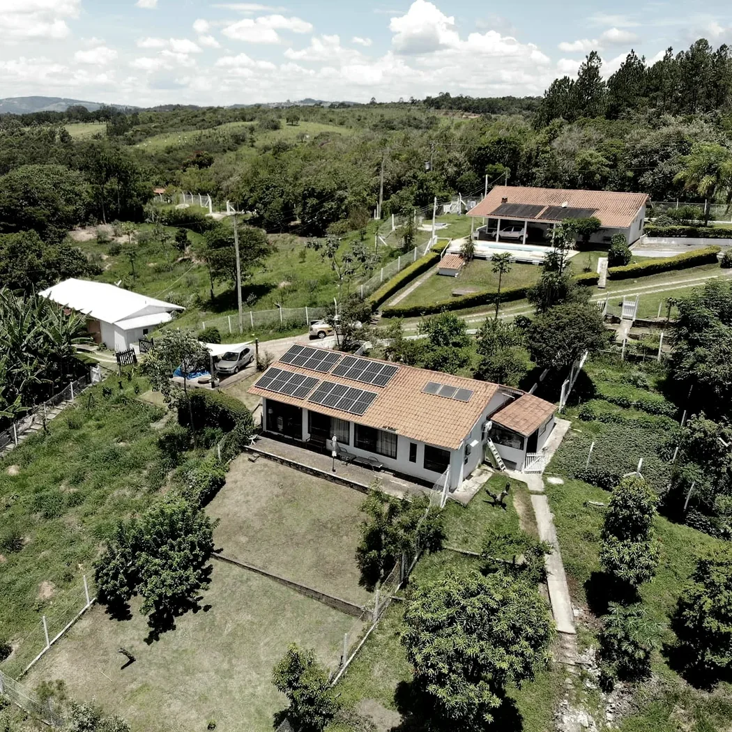 Vista aérea de chácara com casa de telhado colonial e módulos fotovoltaicos, cercada por área verde