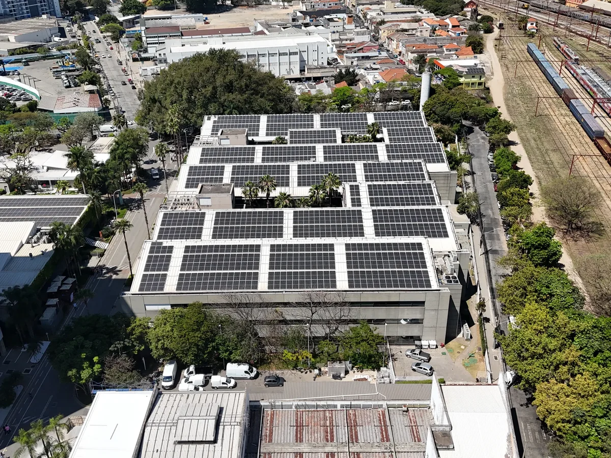 Prédio comercial visto de cima, com a cobertura tomada por módulos fotovoltaicos, cercado por avenida, prédios e linha férrea