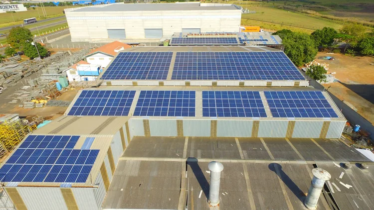 Conjunto de galpões industriais vistos de cima, com fileiras de módulos fotovoltaicos alinhadas nos telhados